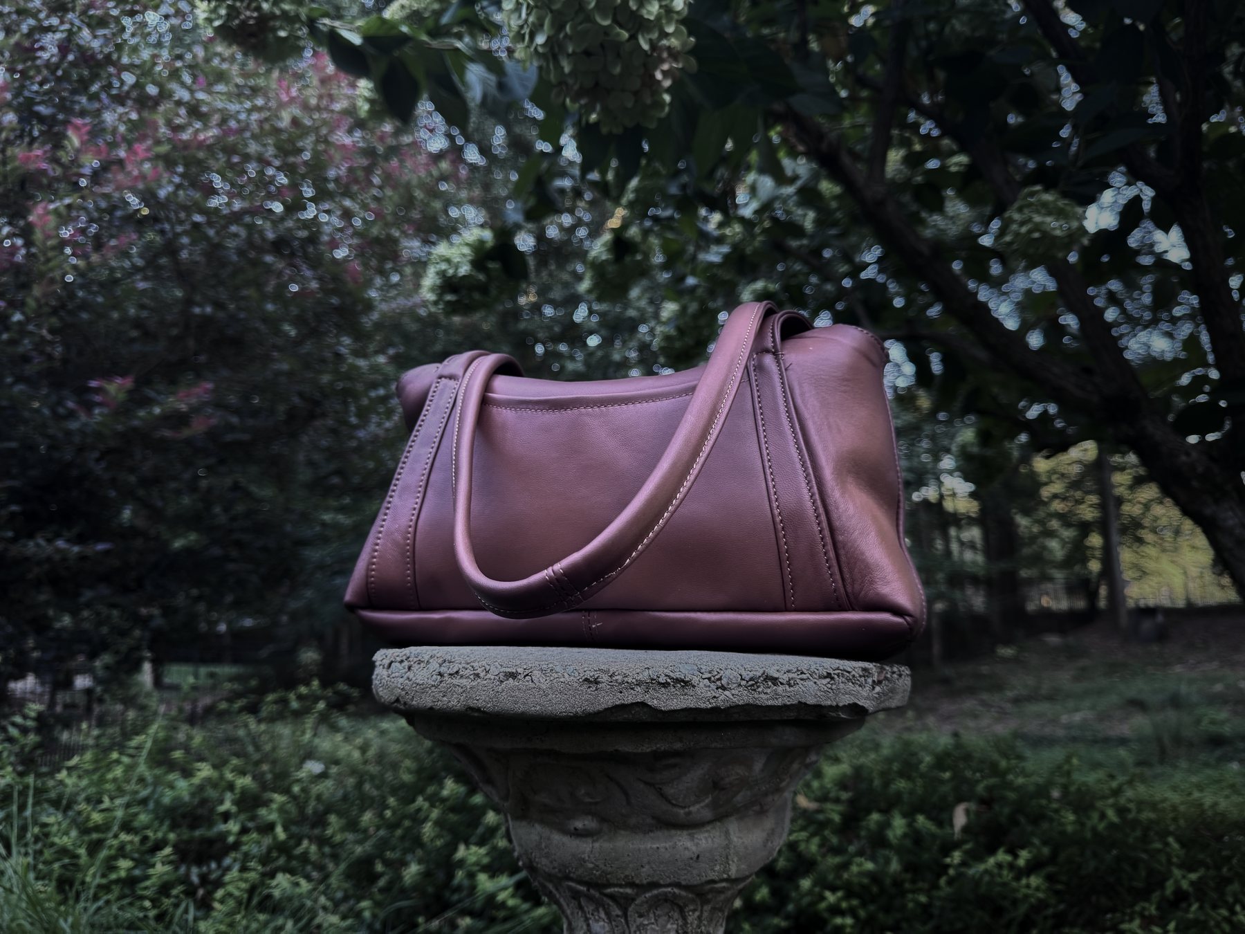 The Oxbo Shoulder in damson lambskin on a stone plinth, lit from the left in a dark garden